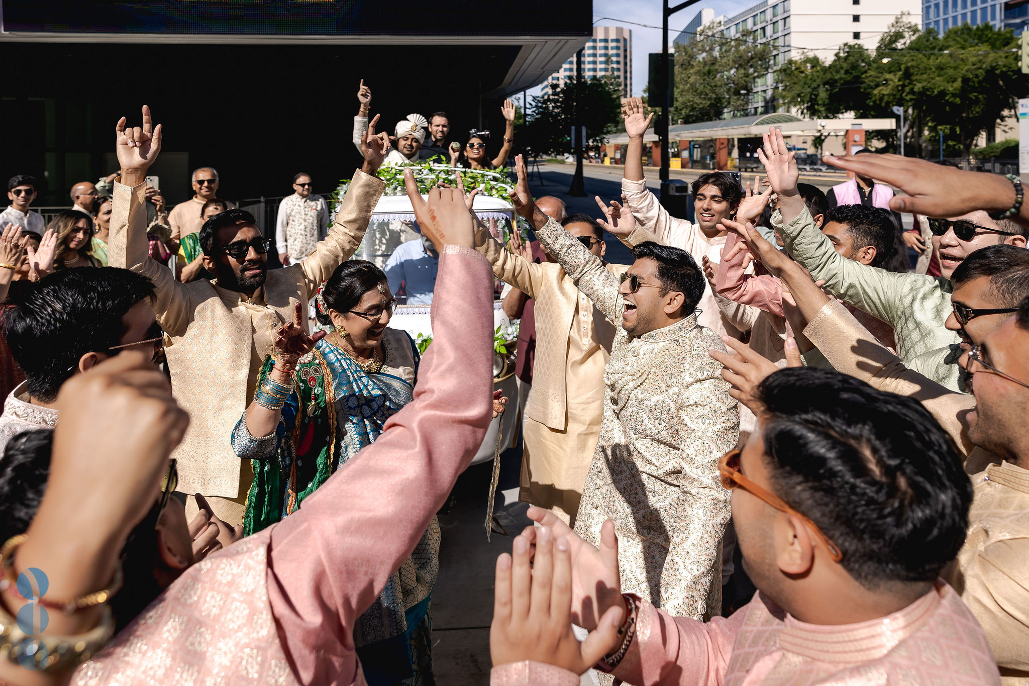 Family and groomsmen dancing with their hands up during a daytime baraat, with the groom in his safa cheering from a flower decorated cart behind them