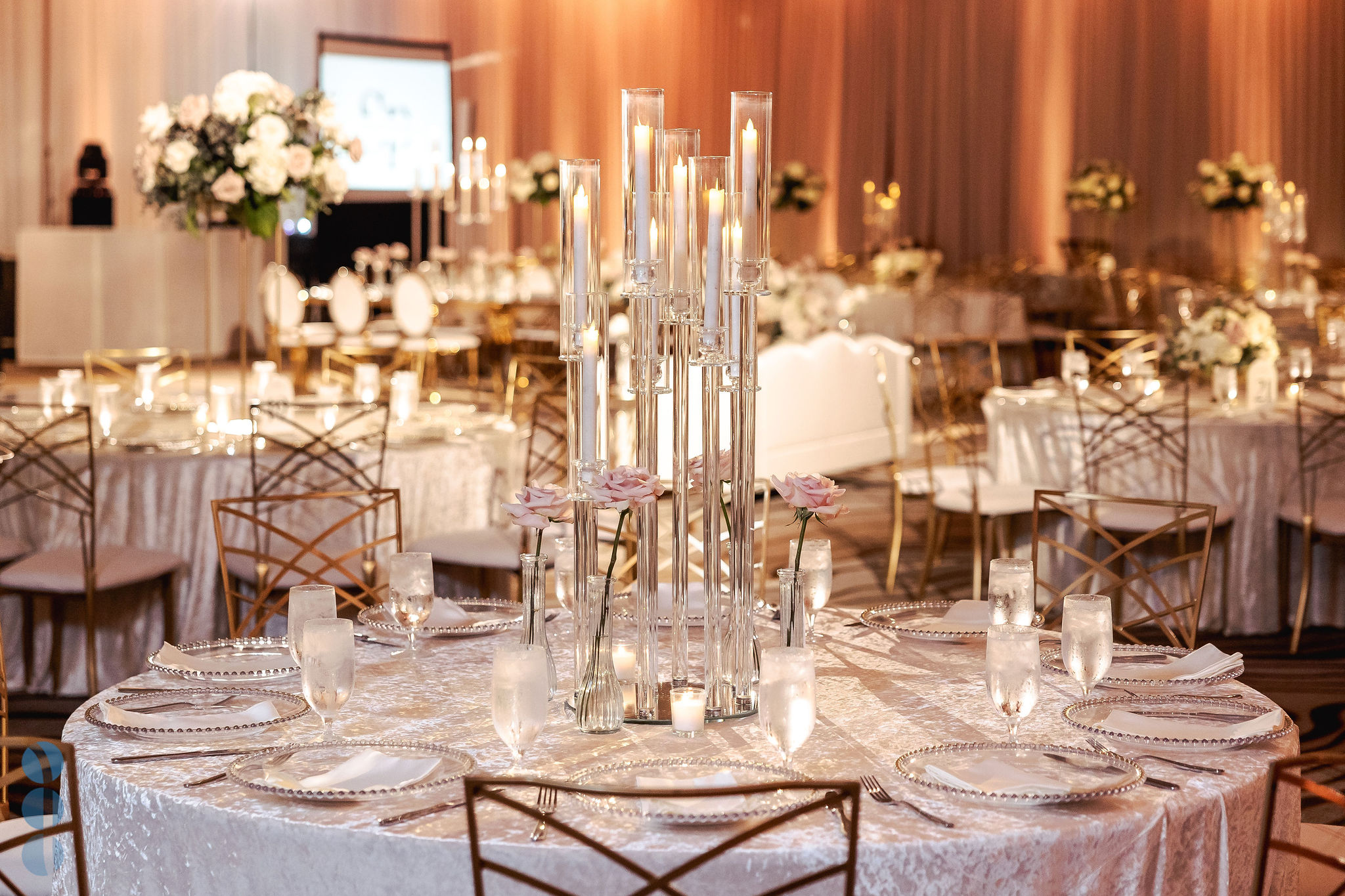Wedding reception tables with tall glass candle centerpieces and blush roses, with the ballroom drapery washed in warm amber uplighting