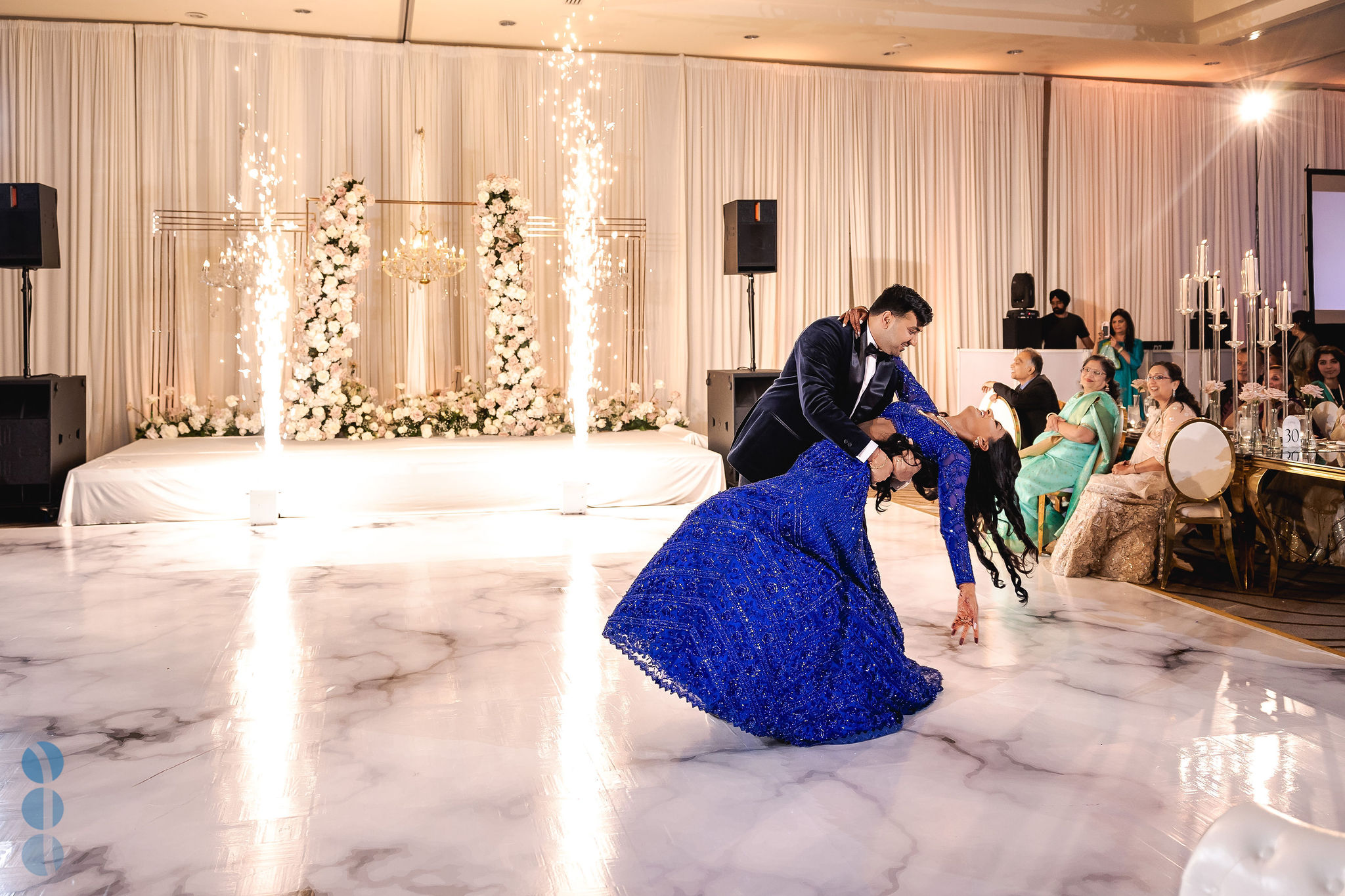 Couple finishing their first dance with a dip on a white marble dance floor as two cold spark fountains fire in front of a floral stage