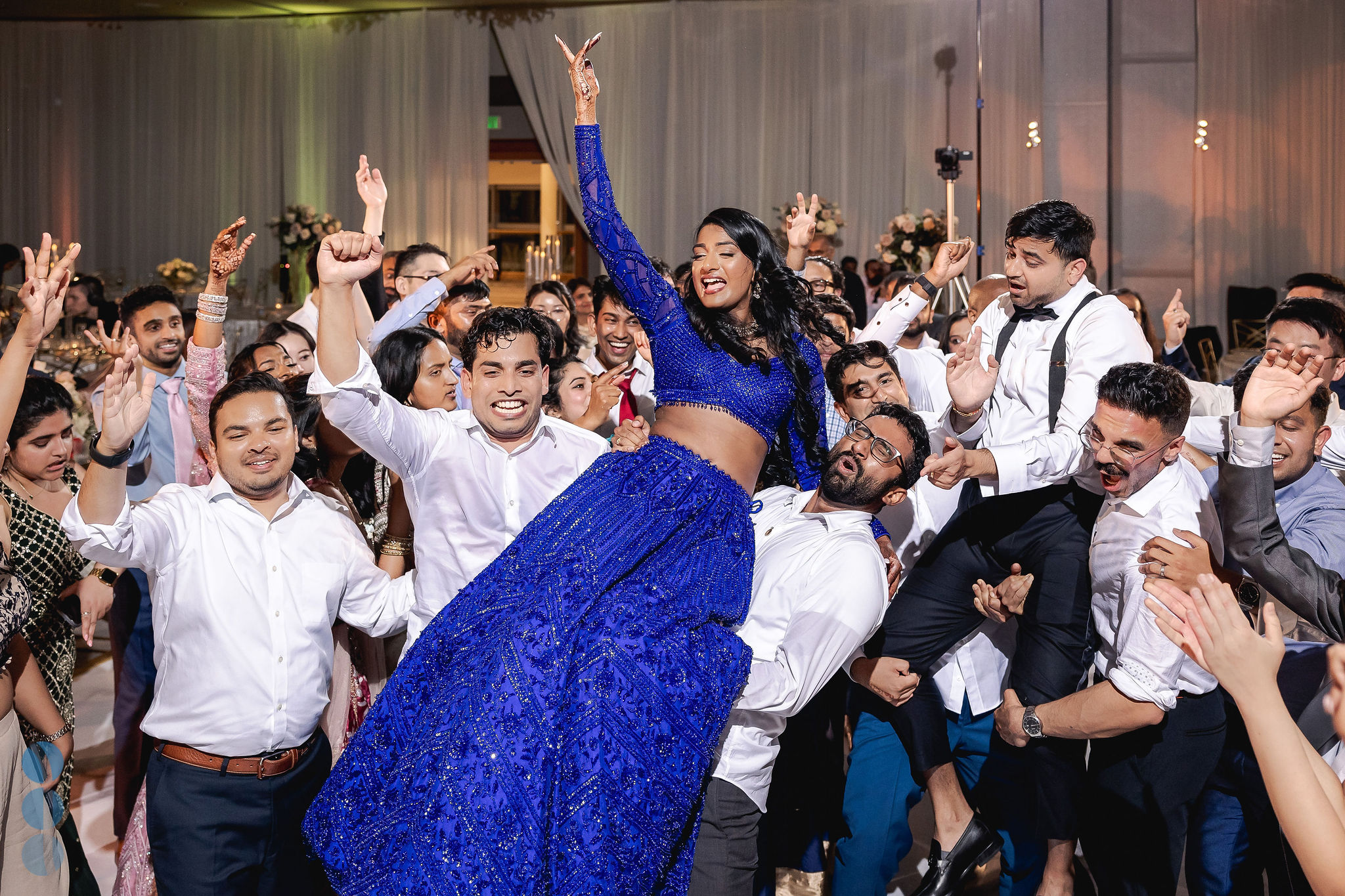 Bride in a royal blue lehenga lifted up by friends on a packed dance floor, dancing with her hand in the air