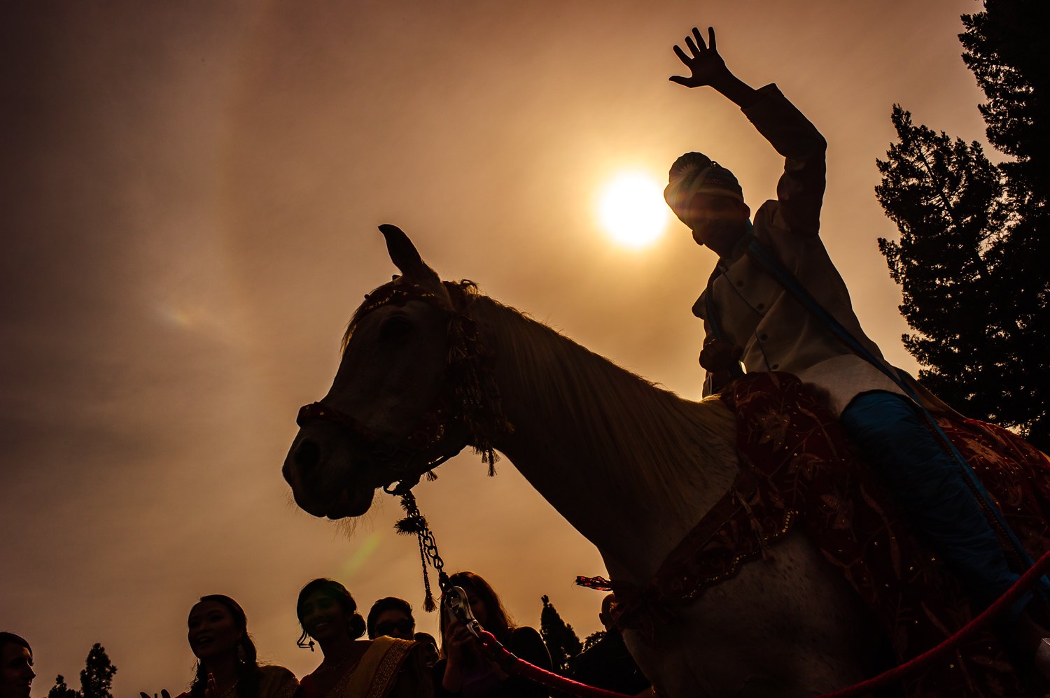 Groom waving from a decorated white horse, silhouetted against a golden sun during his baraat as family dances below