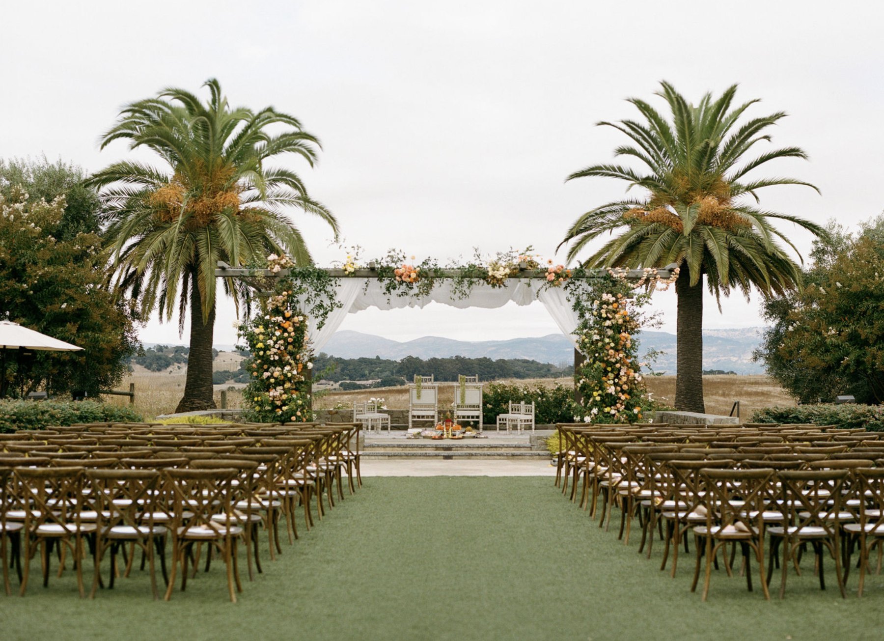 Outdoor Indian wedding ceremony at Carneros Resort in Napa, with rows of guest chairs facing a floral mandap framed by palm trees and hills