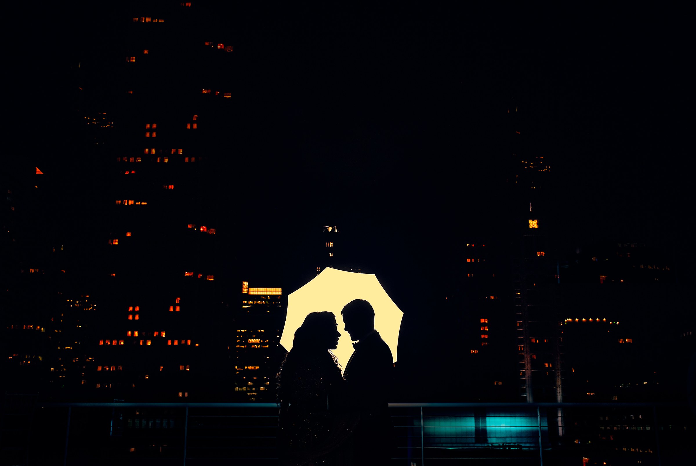Silhouette of a bride and groom under a glowing backlit umbrella against a dark city skyline at night
