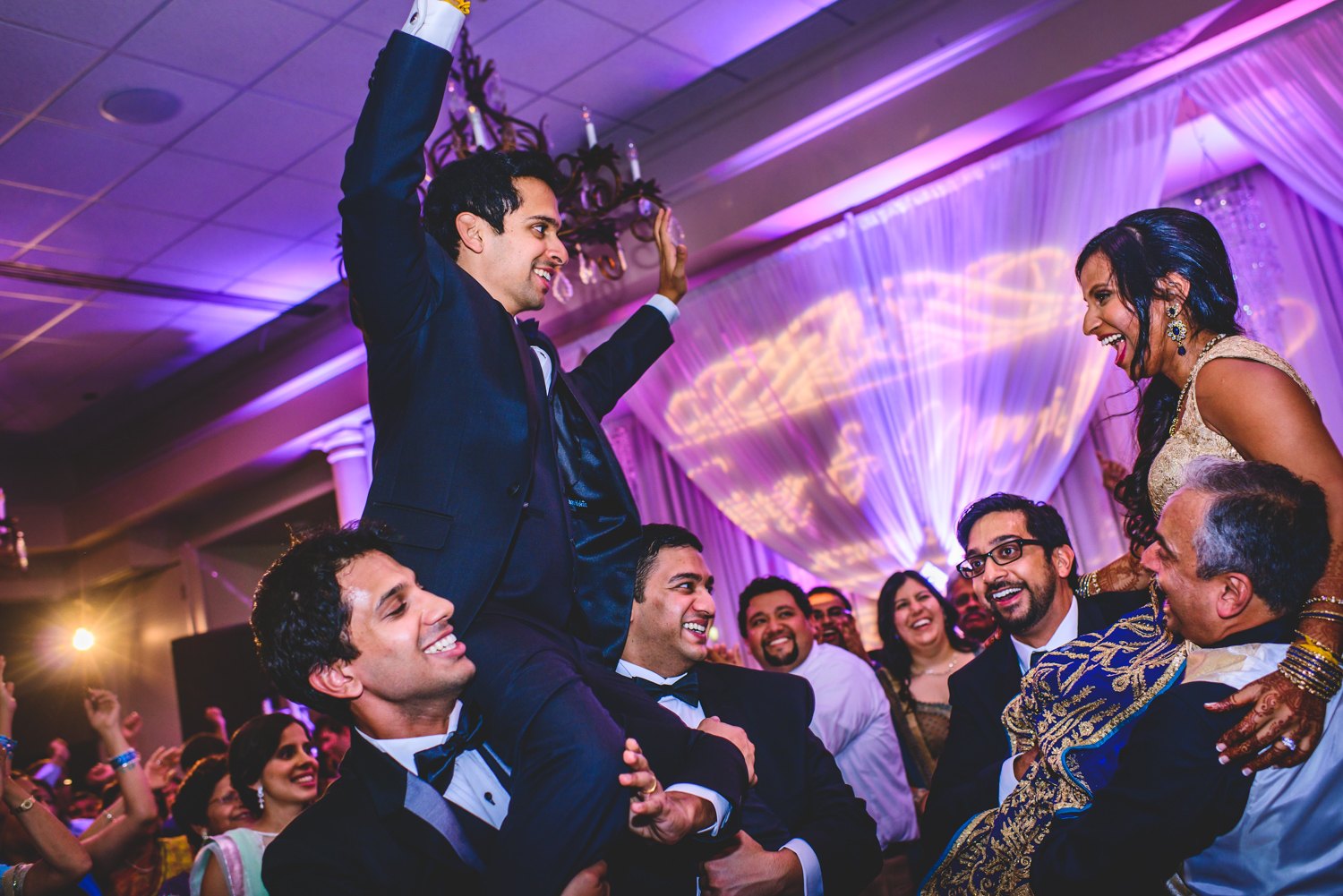 Bride and groom lifted onto shoulders by family on the dance floor, with their custom monogram projected in gold on the purple uplit drapery behind them
