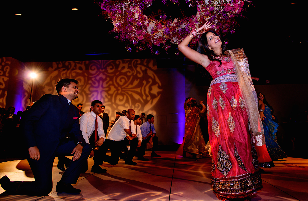 Bride dancing under a hanging floral installation while groomsmen kneel, with gold damask gobo patterns and purple uplighting across the ballroom wall and dance floor