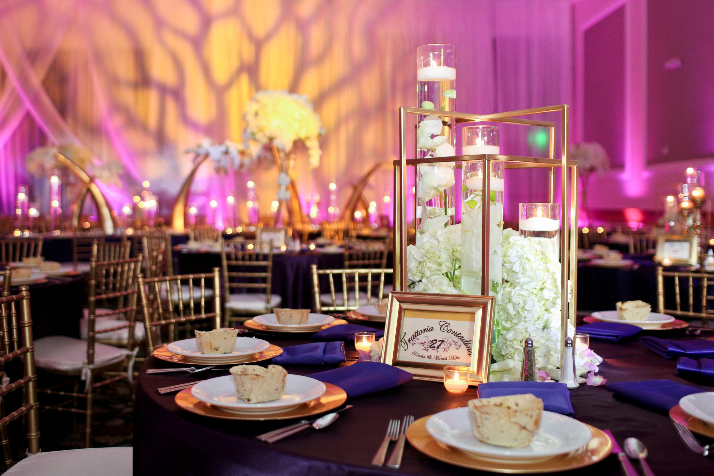 Wedding reception tables with gold centerpieces and floating candles, with an organic gobo pattern in gold across the draped wall and magenta uplighting around the ballroom