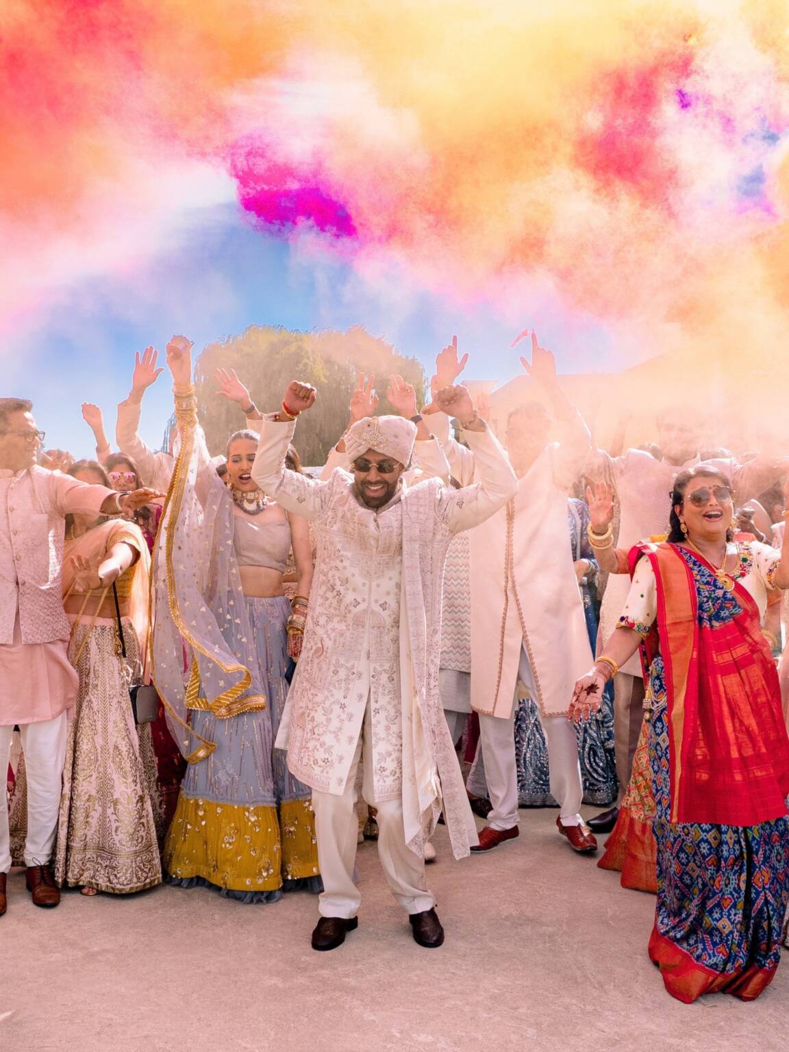 Groom in an ivory sherwani dancing with his hands up at his baraat as family and friends cheer under clouds of pink, orange, and blue color smoke